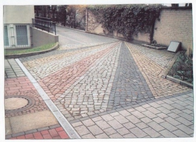 Ułożona nawierzchnia z różnokolorowych kostek, tworząca wzór rozchodzący się promieniście, widoczne fragmenty budynku i zieleni w tle.